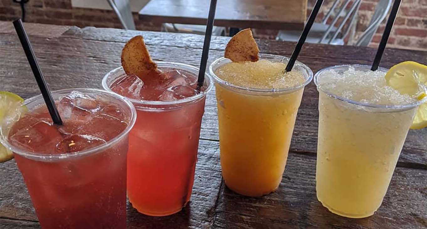 4 cocktails in separate cups with fruit garnish and black straw.