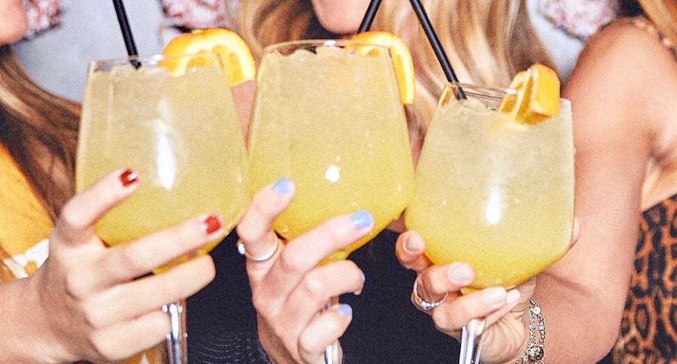 3 women each holding a mimosa garnished with an orange slice and black straw.