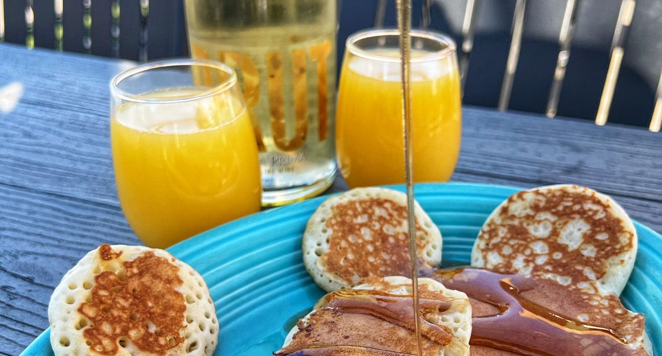 Wooden table with two mimosas and a blue plate holding pancakes with syrup being poured on top.