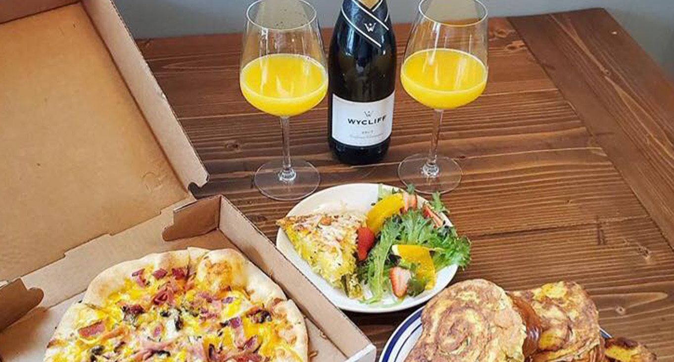 Two mimosas, a bottle of champagne, a box of breakfast pizza, plate of fruit, and various pastries on a wooden table. 